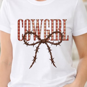 Puede incluir: Camiseta blanca con la palabra "COWGIRL" en un diseño a cuadros rojos y blancos. Un lazo de alambre de púas marrón está centrado debajo del texto, agregando un toque rústico a la camiseta gráfica.
