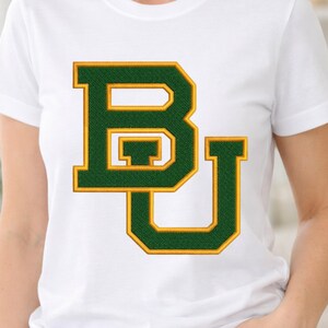 May include: White t-shirt featuring a green and gold embroidered "BU" logo. The letters are outlined in gold and filled with a textured green fabric. The shirt is a classic crew neck style.