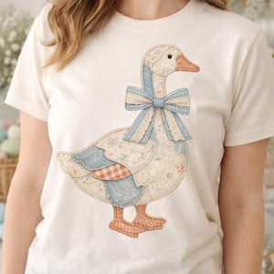 Puede incluir: Camiseta color crema con un diseño caprichoso de ganso. El ganso está adornado con un lazo azul y detalles de patchwork, incluyendo patrones florales y de cuadros. El ganso tiene patas y pico naranjas.