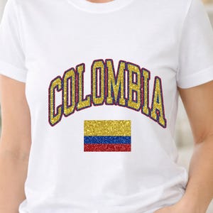 Puede incluir: Una camiseta blanca de manga corta con la palabra COLOMBIA en una fuente arqueada de purpurina dorada con un borde azul oscuro. Debajo del texto hay un gráfico rectangular de la bandera de Colombia en purpurina amarilla, azul y roja.