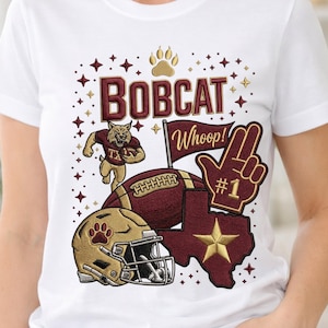 Op de afbeelding: Wit T-shirt met een bordeauxrode en gouden grafische afbeelding. Het ontwerp bevat het woord "BOBCAT", een voetbal, een helm, een pootafdruk, een Texas-omtrek en de zin "Whoop!" met een handgebaar voor nummer één.