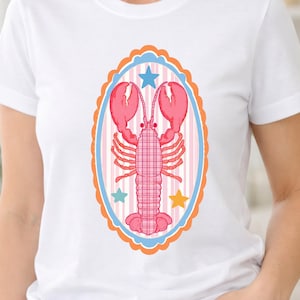 Puede incluir: Una camiseta blanca con un gráfico central de una langosta rosa con un patrón de cuadros en el cuerpo. La langosta está encerrada en un marco ovalado con un borde naranja festoneado, ribete azul claro y rayas verticales rosas, acentuado por tres estrellas.