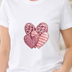 Puede incluir: Una camiseta blanca de manga corta con un gráfico central de tres corazones superpuestos. Dos corazones muestran un estampado de leopardo en rosa y rojo oscuro, mientras que el tercer corazón presenta rayas horizontales rosas y rojas brillantes.