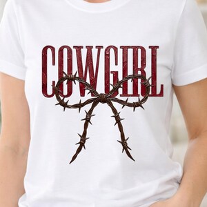 Puede incluir: Camiseta blanca con la palabra "COWGIRL" en fuente burdeos texturizada. Debajo, un diseño de lazo de alambre de púas marrón. Es una prenda informal.