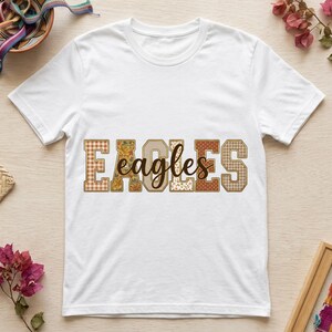 Puede incluir: Una camiseta blanca de manga corta con la palabra EAGLES en letras grandes de bloque con patrones variados en tonos marrón, beige y naranja, superpuesta con la palabra eagles en una fuente cursiva marrón oscuro en el centro.