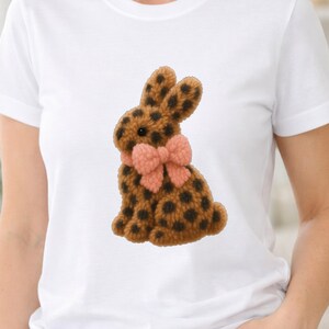 May include: White t-shirt with a brown bunny design. The bunny has black spots and a pink bow. The design is textured, giving a plush appearance. The t-shirt is a classic, casual style.