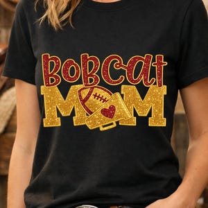 Puede incluir: Camiseta negra con un diseño de purpurina dorada y roja. El diseño presenta las palabras "Bobcat Mom" con un balón de fútbol americano y un megáfono. La camiseta es de material suave.