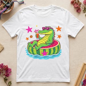 Puede incluir: Una camiseta blanca con un gráfico de un cocodrilo verde con gafas de sol rosas, relajándose en un flotador con forma de sandía sobre agua azul. El cocodrilo sostiene una bebida naranja con una sombrilla rosa, rodeado de estrellas naranjas y rosas.
