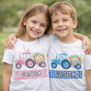 Puede incluir: Dos niños con camisetas blancas con diseños de tractores. Una camiseta presenta un tractor rosa, la otra un tractor azul, ambos tirando de un carro de huevos de Pascua. Cada camiseta tiene una pancarta de texto personalizada.