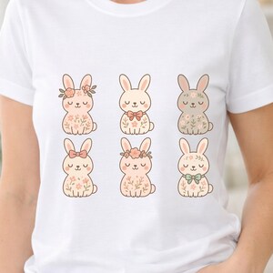 Puede incluir: Camiseta blanca con seis conejitos de dibujos animados con detalles florales y lazos. Los conejitos son de varios colores, incluyendo rosa, beige y gris. El diseño tiene un tema lindo y caprichoso.