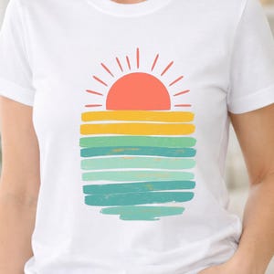 Peut inclure: Un t-shirt blanc à manches courtes arborant un motif central représentant un soleil stylisé se couchant à l'horizon. Le design se compose d'un demi-cercle corail avec des rayons, suivi de bandes horizontales aux traits de pinceau jaunes et verts.