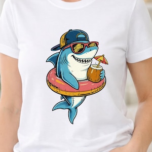 Puede incluir: Una camiseta blanca con un gráfico de un tiburón azul que lleva una gorra azul y amarilla y gafas de sol rojas. El tiburón está dentro de un flotador rosa y amarillo, sosteniendo una bebida de coco con una sombrilla y el texto SUMMER VIBES.