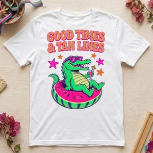 Puede incluir: Una camiseta blanca con un gráfico de un caimán verde con gafas de sol moradas, descansando en un flotador de sandía mientras sostiene un cóctel con sombrilla. El texto GOOD TIMES & TAN LINES aparece arriba en color naranja.