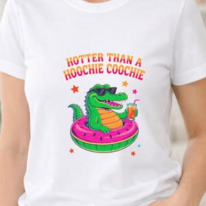 May include: A white short-sleeved t-shirt featuring a graphic of a green alligator wearing sunglasses, floating in a pink watermelon-patterned inner tube while holding a drink. The text above reads HOTTER THAN A HOOCHIE COOCHIE in orange and pink.