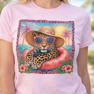 Puede incluir: Camiseta rosa claro con un estampado gráfico de un leopardo con gafas de sol y sombrero de paja, relajándose en una boya rosa. El diseño incluye una escena de playa tropical con palmeras y una puesta de sol.