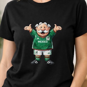 May include: A black short-sleeved t-shirt featuring a graphic of a cartoon character with a white mustache wearing a green and white Mexico soccer jersey, white shorts, green socks, and black cleats with arms outstretched in a welcoming gesture.
