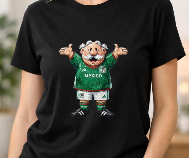 May include: A black short-sleeved t-shirt featuring a graphic of a cartoon character with a white mustache wearing a green and white Mexico soccer jersey, white shorts, green socks, and black cleats with arms outstretched in a welcoming gesture.