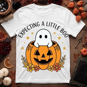 Puede incluir: Una camiseta blanca de manga corta con un gráfico de un fantasma blanco sentado dentro de una calabaza naranja, rodeado de estrellas amarillas y flores de otoño, con el texto negro "EXPECTING A LITTLE BOO" impreso encima.