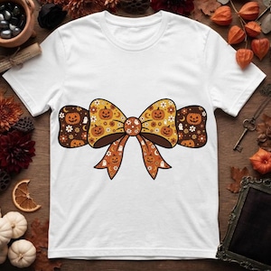 Puede incluir: Una camiseta blanca de manga corta con un gran gráfico central en forma de lazo. El lazo está decorado con un patrón repetitivo de calabazas de Halloween naranjas, pequeños fantasmas blancos, flores blancas y lunas crecientes sobre un fondo marrón.