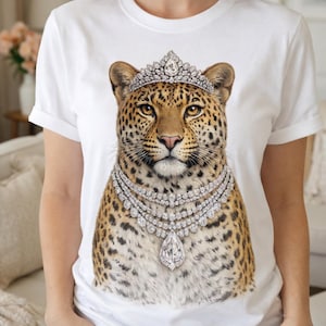 Puede incluir: Camiseta blanca con una ilustración de un leopardo adornado con una tiara y un collar de diamantes. El leopardo tiene un pelaje manchado y es el elemento central del diseño. El collar presenta un gran colgante en forma de pera.