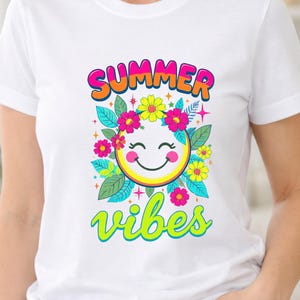 Puede incluir: Una camiseta blanca con un estampado gráfico colorido. El diseño incluye el texto "SUMMER" en naranja y rosa, y "vibes" en verde lima. Una cara sonriente amarilla central está rodeada de flores rosas y amarillas y hojas de color azul verdoso.