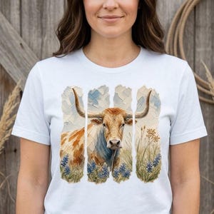 Puede incluir: Una camiseta blanca de manga corta con un gráfico central de una vaca longhorn en un campo de flores azules. La ilustración se presenta en tres paneles verticales con un estilo pictórico texturizado, mostrando el pelaje marrón y blanco.