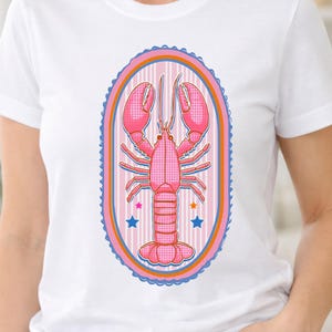 Puede incluir: Una camiseta blanca de manga corta con un gráfico central de una langosta rosa con un patrón de cuadrícula. La langosta está encerrada en un marco ovalado con rayas rosas y naranjas, un borde azul festoneado y pequeñas estrellas azules y naranjas.