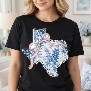 Puede incluir: Camiseta negra con un diseño del estado de Texas. El contorno del estado está relleno con un estampado floral azul y una cinta rosa. El diseño está centrado en la parte delantera de la camiseta.