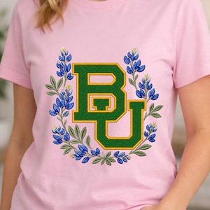 Puede incluir: Camiseta rosa claro con el logotipo "BU" verde y dorado, rodeado por una corona de flores de altramuces y hojas verdes. El diseño está bordado en la parte delantera, creando un efecto texturizado. La camiseta tiene mangas cortas.