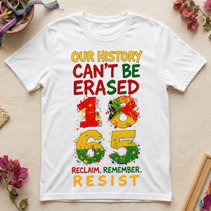 Könnte beinhalten: Ein weißes Kurzarm-T-Shirt mit dem Text "OUR HISTORY CAN'T BE ERASED" in Gelb, Rot und Grün. Darunter ist die Zahl "1865" in großen, farbspritzigen Ziffern abgebildet, gefolgt von "RECLAIM. REMEMBER. RESIST." in Rot, Grün und Gelb.