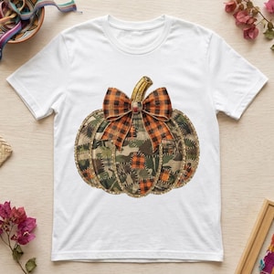 Puede incluir: Una camiseta blanca de manga corta con un gráfico central de una calabaza hecha de un mosaico de patrones de tela de camuflaje y cuadros naranjas. La calabaza está rematada con un gran lazo de cuadros naranjas y negros y un tallo dorado.
