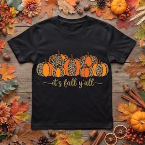 Puede incluir: Una camiseta negra de manga corta con un gráfico de seis calabazas naranjas con estampado de leopardo. Debajo de las calabazas, el texto "it's fall y'all" está impreso en una fuente cursiva dorada en el centro de la camiseta.