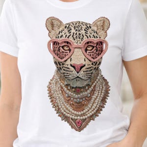 Puede incluir: Camiseta blanca con un leopardo bordado que lleva gafas rosas en forma de corazón y un collar de perlas. El leopardo tiene manchas marrones y negras. La camiseta es de cuello redondo.