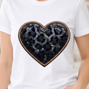 Puede incluir: Camiseta blanca con un parche grande en forma de corazón. El parche tiene un centro de lentejuelas azul oscuro y un borde dorado. Una prenda casual y cotidiana.