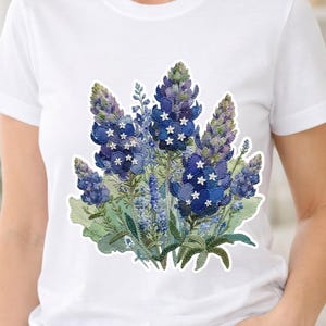 Puede incluir: Camiseta blanca con un diseño floral de altramuces azules. El diseño presenta flores azules y moradas con acentos de estrellas blancas, sobre hojas verdes. El gráfico es una aplicación tipo parche.