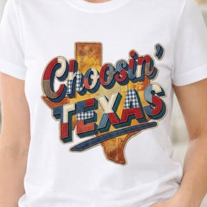 Puede incluir: Camiseta blanca con la frase "Choosin' Texas" en un diseño colorido estilo patchwork. El diseño incorpora la forma del estado de Texas, con detalles en rojo, azul y marrón. La camiseta está hecha de un material suave y cómodo.