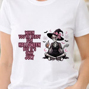 Puede incluir: Una camiseta blanca con un gráfico de un esqueleto vestido de bruja, con gafas en forma de estrella y sosteniendo una bebida boba. El texto dice: WHEN YOU'RE READY FOR HALLOWEEN BUT IT'S ONLY JULY. El diseño incluye murciélagos, telarañas y lunas.