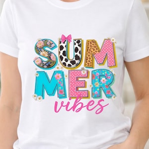 Puede incluir: Una camiseta blanca con el texto SUMMER VIBES en letras grandes con diversos patrones. Las letras incluyen diseños florales, manchas de vaca, estampado de leopardo y lunares rosas, decoradas con un lazo rosa y margaritas blancas.