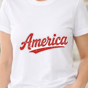 Puede incluir: Una camiseta blanca de manga corta con la palabra America impresa en el pecho en una fuente roja de estilo manuscrito desgastado, con un contorno blanco y un subrayado rojo que se extiende desde la última letra.