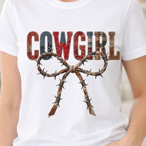 Puede incluir: Camiseta blanca con la palabra "COWGIRL" en un diseño de patchwork con patrones rojos, azules y marrones. Un lazo de alambre de púas está centrado debajo del texto, agregando un toque rústico al diseño.