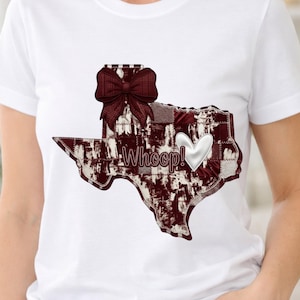 Puede incluir: Camiseta blanca con un diseño en forma de Texas en burdeos y blanco, con un lazo y un corazón. El diseño incluye la palabra "Whoop!" en blanco. La camiseta está hecha de un material suave y cómodo.