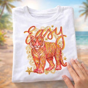 Puede incluir: Camiseta blanca con una ilustración de un tigre en naranja, amarillo y rosa. La palabra "Easy" está escrita encima del tigre en una fuente cursiva. Estrellas doradas y un borde decorativo rodean el diseño del tigre.
