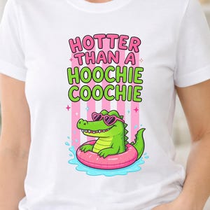 Puede incluir: Una camiseta blanca con un gráfico de un caimán verde con gafas de sol rosas en forma de corazón, flotando en un flotador rosa. Sobre el caimán, el texto dice HOTTER THAN A HOOCHIE COOCHIE en letras de bloque de color rosa y verde.