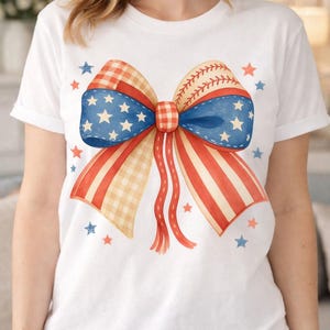 Puede incluir: Camiseta blanca con un diseño de lazo patriótico. El lazo incorpora elementos de la bandera estadounidense y costuras de béisbol, con colores rojo, blanco y azul. Estrellas dispersas completan el look festivo.