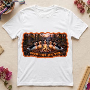 Puede incluir: Una camiseta blanca de manga corta con un gráfico rectangular de borde naranja. La imagen muestra cinco patos blancos con sombreros de bruja y capas negras alrededor de una fogata en un bosque. El texto dice SUPPORT YOUR LOCAL COVEN.