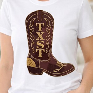 Op de afbeelding: Wit T-shirt met een bruin en goud cowboylaarsontwerp. De laars heeft de gouden letters "TXST". Het ontwerp is gedetailleerd met stiksels en decoratieve elementen, wat een rustieke en stijlvolle uitstraling creëert.