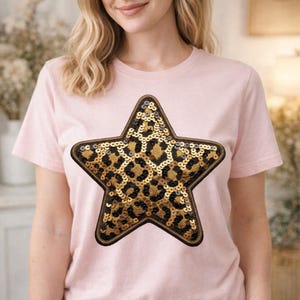Puede incluir: Camiseta rosa claro con un diseño de lentejuelas en forma de estrella. La estrella presenta un estampado de leopardo en lentejuelas doradas y negras, delineado con un borde marrón. La camiseta es de manga corta.