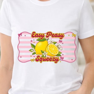 Puede incluir: Una camiseta blanca de manga corta con un gráfico central de dos limones amarillos con hojas verdes y flores blancas. El diseño incluye un fondo de rayas rosas y blancas con el texto "Easy Peasy Squeezy" escrito en letras amarillas.