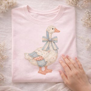 Puede incluir: Una sudadera rosa claro con un diseño de ganso aplicado. El ganso tiene un lazo azul, alas con estampado floral y un parche a cuadros rojos y blancos. El diseño está centrado en la parte delantera de la camisa.