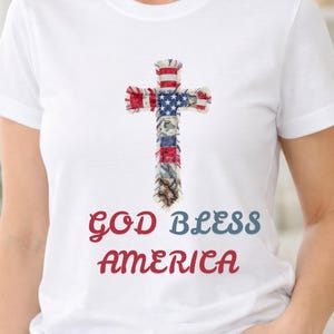 Puede incluir: Camiseta blanca con un diseño de cruz que incorpora los colores de la bandera estadounidense. La cruz está hecha de tiras de tela. Las palabras "GOD BLESS AMERICA" están impresas en rojo y azul.